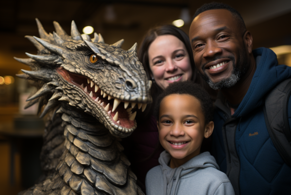 A man and a woman are posing for a picture with a dragon statue creature in what appears to be a museum setting. The man, who is around 34 years old, is wearing a tie and has a big smile on his face. The woman, who is around 30 years old, is also smiling. In the background, there is a young boy, around 11 years old, smiling as well. The scene includes a desk in the background. The primary colors in the image are shades of brown and black, with accents of a lighter brown. The overall mood of the image is joyful and family-oriented
