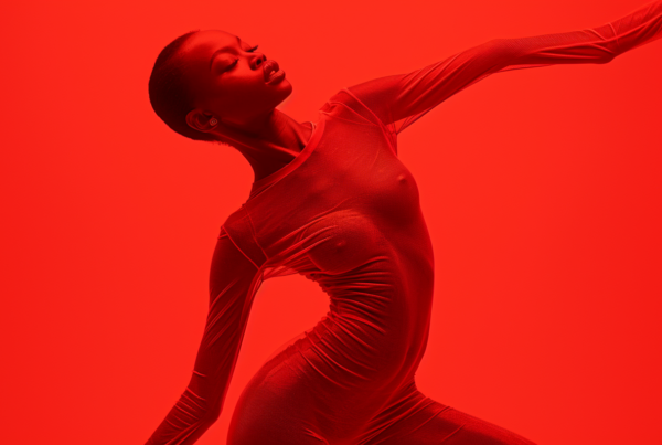 In this image, a woman is seen wearing a vibrant red bodysuit, striking a pose in front of a red background. She exudes confidence and grace as she poses for the camera, with her arms outstretched. The bold red colors of the bodysuit and background create a striking visual contrast, drawing attention to the woman''s figure and movement. The woman''s posture suggests she may be dancing or performing, adding a dynamic element to the composition. Her expression is focused and determined, adding to the sense of energy and motion captured in the photograph.