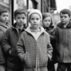 A black and white photo depicting a group of children standing next to each other. The children are a mix of boys and girls, with some wearing coats and jeans, and one girl wearing a dress. The image shows a total of eight children, with some wearing hats. The children are standing in front of what appears to be a store, as there is a window in the background. The photo captures a candid moment of the children interacting with each other. The children range in age from 5 to 9 years old, and there is a mix of genders present.