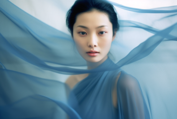 A young woman with a serene expression is captured in the image, wearing a beautiful blue dress that catches the eye. Her gaze is direct and engaging, as if she is looking straight at the camera. The woman''s face is partially covered by a delicate veil, adding an air of mystery and elegance to her appearance. The colors in the image are predominantly shades of blue and gray, creating a soothing and harmonious visual aesthetic. Overall, the scene conveys a sense of grace and sophistication, making it a captivating and intriguing portrait.