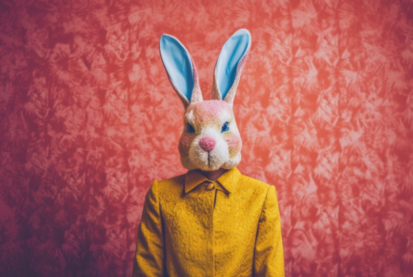 A person is wearing a bunny mask on their head, with a stuffed toy bunny visible in the background. The stuffed toy is brown with a yellow coat, and is placed against a pink background. The person is not facing the camera directly, but the bunny mask is prominently displayed on their head. The colors in the image include shades of brown, yellow, red, and a hint of blue. The overall composition gives a playful and whimsical feel, perfect for Easter or a costume party. This image could be used for a dataset on bunny-themed accessories or for creative mask designs.