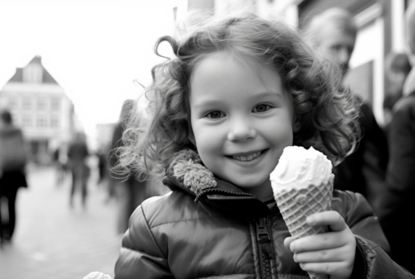 In this black and white image, a little girl is joyfully holding a large ice cream cone in her hand. She is smiling brightly as she enjoys her sweet treat. The background is slightly blurred, focusing all attention on the girl and her ice cream. Another person can be seen in the background, also holding an ice cream cone. The girl is wearing casual clothing and seems to be in a happy and carefree moment. The image captures a simple yet delightful scene of childhood innocence and happiness.