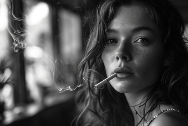 In this black and white photo, a young woman is depicted smoking a cigarette. The woman, who appears to be around 23 years old, has long hair and is wearing a necklace. The image focuses on her face, capturing her holding the cigarette in her mouth. In the background, there is a small flower in a vase. The woman''s expression is neutral as she smokes. The overall tone of the image is nostalgic and vintage. The contrast between the dark tones and the woman''s white skin adds to the dramatic effect of the composition.