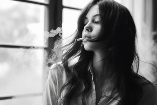 In this black and white photo, a woman is seen standing in front of a window, smoking a cigarette. The woman has long black hair and is wearing a simple outfit. The photo captures her profile as she takes a drag from the cigarette, with the smoke swirling around her. The room behind her is dimly lit, emphasizing her silhouette against the window. The contrast between light and dark adds a dramatic effect to the image. The overall mood is contemplative and mysterious, making the viewer wonder about the woman''s thoughts and emotions.