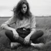 A serene scene captured in black and white, where a young woman is sitting gracefully in a vast field of tall grass. Her long, flowing hair dances in the wind as she sits with her legs crossed, embodying a sense of peace and tranquility. She is wearing a pair of boots and a belt, adding a touch of ruggedness to her otherwise ethereal presence. In the background, the grass sways gently, creating a harmonious atmosphere. The image exudes a sense of calm and connection with nature, inviting the viewer to pause and appreciate the beauty of the moment