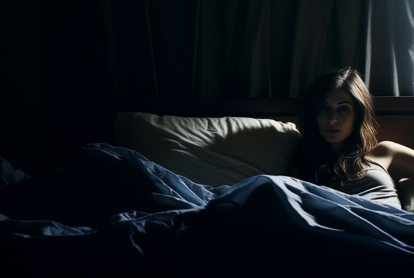 In the image, a young woman is peacefully laying in bed with a cozy blue blanket covering her. The room is dimly lit, creating a serene and relaxing atmosphere. A fluffy pillow is placed next to her, adding to the comfort of the scene. The woman appears to be in her early twenties and is the focus of the image, exuding a sense of tranquility and relaxation. The overall color palette of the image consists of shades of blue, gray, and white, enhancing the peaceful and calming vibe of the setting
