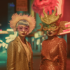 Two women are showcased in striking gold outfits on a lively city street. The first woman captivates in a shimmering gold dress and matching mask, while the second woman dons a gold jacket and pink mask. Additionally, she sports a pink feathered hat that adds a touch of elegance to her ensemble. Both women exude confidence and style as they stand gracefully against the urban backdrop. The women''s vibrant costumes and accessories create a visually stunning scene, drawing attention to their fashionable and bold choices in attire.