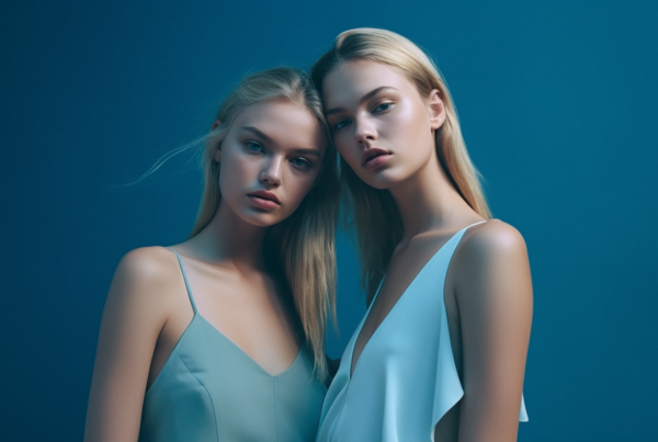 In this image, we see two young women wearing elegant blue dresses. Both women are posing for a picture, standing confidently with one hand on their hips. The woman on the left has long blonde hair and blue eyes, while the woman on the right has dark hair. They are both models, exuding grace and style in their attire. The background is a solid blue color, emphasizing the beauty of the women and their outfits. The women are the focal point of the image, radiating confidence and sophistication. This picture captures a moment of glamour and poise, perfect for a fashion magazine or advertisement.