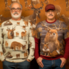 Two men are standing next to each other in front of a wall that is adorned with intricate designs of deer and bears. The men are wearing matching sweaters, and one of them is wearing a hat and glasses. The wall art features various images of deer and bears in different poses. The men appear to be admiring the artwork on the wall. One man is older, around 70 years old, while the other is younger, around 58 years old. The colors in the scene are predominantly earthy tones, with browns and oranges dominating the color palette.