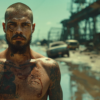 A man with tattoos on his chest is standing in a dirty area, surrounded by a car and a van. The man appears to be muscular and has a beard. The tattoos on his chest are visible, adding to his rugged appearance. The area he is in looks gritty and urban, with debris scattered around. The car and van in the background are slightly blurry, indicating movement in the scene. The man exudes a sense of toughness and confidence as he stands in this rough environment. The overall atmosphere is one of urban decay and edginess.