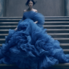 A young woman with long brown hair is seated gracefully on a grand staircase, wearing a stunning blue gown. The gown flows elegantly around her, creating a striking contrast against the neutral stone of the stairs. The woman exudes poise and elegance as she gazes off into the distance, lost in thought. The intricate details of the gown''s design are highlighted by the soft lighting, making it appear even more regal. The scene conveys a sense of beauty and sophistication, with the woman serving as a captivating focal point in this architectural setting.