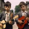 Two young boys, around 7 and 9 years old, are dressed in suits and ties, standing in front of a house. Both boys are holding guitars, one with a brown guitar and the other with a black guitar. The younger boy has short brown hair and is on the left side, while the older boy with longer hair is on the right. The house behind them has a window, and the scene seems to be in a residential area. The boys look confident and focused as they hold their guitars, ready to play music together.