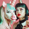In this vibrant and whimsical image, two women are seen wearing elaborate unicorn makeup, complete with colorful horns and sparkles. The women are standing next to a beautifully decorated birthday cake, adorned with candles. The cake is a stunning display of pastel colors, with pink and white frosting. One of the women is looking directly at the camera, showcasing her intricate makeup, while the other woman is admiring the cake in the background. The overall scene exudes joy and celebration, with a magical and fantastical atmosphere. The image captures a moment of fun and festivity, perfect for a birthday party or special occasion.