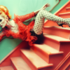 A woman is sitting on a staircase wearing a red dress and polka dot tights. She is accompanied by a bottle of wine with a red ribbon around it. The woman is the focus of the image, with her outfit standing out against the stairs. In the background, there is a dalmatian dog figurine with a red background, adding a playful element to the scene. The woman''s outfit and the dalmatian dog create a whimsical and fashionable atmosphere. The overall composition is colorful and eye-catching, perfect for a fashion or lifestyle theme.
