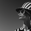 In this black and white photo, a woman is depicted wearing a stylish hat and sunglasses. The woman, who appears to be a model, is dressed in a striped suit and is confidently posing for the camera. The hat she is wearing is white with a black band, adding a chic touch to her outfit. The sunglasses she has on are large and black, shielding her eyes from the sun. The image captures her in a moment of elegance and sophistication, making her the epitome of fashion and style. The overall aesthetic is classic and timeless, reminiscent of vintage glamour.