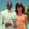 A man and a woman are dressed in a stylish white suit with sunglasses. The woman, who appears to be in her late 20s, is wearing a peach dress and is holding a small dog in her arms. The man is wearing a blue tie and a white shirt. The image also features a dog with a collar around its neck. The couple is standing together in what seems to be a desert setting. The woman has long hair and is striking a pose while holding the dog. The man is wearing glasses and both individuals are looking fashionable and elegant.