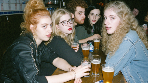 A group of people is gathered around a table, enjoying drinks together. The scene includes a diverse mix of individuals, including a man and several women. One woman stands out with her red hair and glasses, holding a beer. Another woman is wearing a black shirt and red lipstick, also holding a beer. A man with a beard is seated at the table as well. The atmosphere appears relaxed and social, with conversations and laughter likely filling the air. The background shows a bar setting with shelves and cabinets visible. Overall, it''s a casual and lively gathering of friends enjoying each other''s company over drinks.