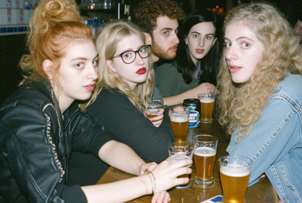 A group of people is gathered around a table, enjoying drinks together. The scene includes a diverse mix of individuals, including a man and several women. One woman stands out with her red hair and glasses, holding a beer. Another woman is wearing a black shirt and red lipstick, also holding a beer. A man with a beard is seated at the table as well. The atmosphere appears relaxed and social, with conversations and laughter likely filling the air. The background shows a bar setting with shelves and cabinets visible. Overall, it''s a casual and lively gathering of friends enjoying each other''s company over drinks.
