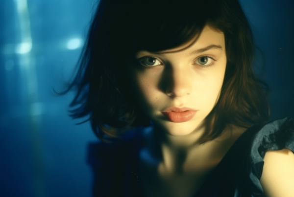 A close-up image of a woman with a blue background, standing in front of a palm tree. The woman is wearing a dark dress and has a serene expression on her face. Her eyes are the focal point of the image, exuding a sense of mystery and elegance. The lighting creates a subtle shadow on her face, adding depth to the composition. The overall color scheme consists of shades of blue, with accents of gold and black. The woman appears to be in her twenties, with a feminine appearance. This image captures a moment of beauty and tranquility.