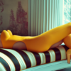 A woman in a yellow outfit is seen laying on a stylish striped couch. She is wearing high heels, with one of them visible in the foreground and another one slightly blurred in the background. The woman''s face is not visible in the image, but she appears to be relaxed and comfortable. The colors in the image are primarily shades of yellow, red, and blue. The overall aesthetic is stylish and modern, with a touch of sophistication. The setting suggests a cozy and chic living room environment.