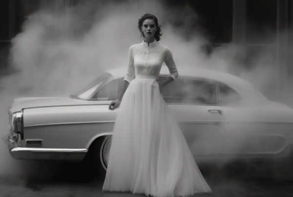 A stunning image captured in black and white featuring a woman in a beautiful white wedding dress standing elegantly next to a sleek car. The woman appears to be the focal point of the image, exuding grace and poise. She is standing confidently with one hand on her hip, showcasing the intricate details of her gown. In the background, a car is visible adding a touch of modernity to the classic scene. The overall composition exudes a sense of sophistication and elegance, making it a timeless and captivating moment.