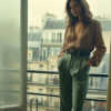 A young woman is standing in front of a window in a hotel room, looking out at a city view. She is wearing a tan shirt and green pants, accessorized with a belt. Her dark hair falls loosely around her shoulders as she gazes outside. The room is decorated in earthy tones, with a cozy ambiance. The woman appears relaxed and contemplative, enjoying the urban scenery from her vantage point. The image captures a moment of quiet reflection, with the woman''s outfit and the room''s decor creating a harmonious composition.
