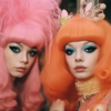 In this image, there are two women with striking pink hair and enchanting blue eyes. The first woman, located on the left side, has pink hair styled in a wig, wearing a black dress. Her eyes are a mesmerizing blue color. The second woman, on the right side, also has pink hair and blue eyes, wearing a pink sweater. Both women exude a unique and vibrant style with their colorful hair and eyes. The image captures their individuality and confidence as they stand out with their unconventional appearanc