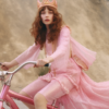 A young woman, approximately 22 years old, is elegantly dressed in a pink dress and a crown on her head. She is confidently riding a pink bicycle in this charming scene. The woman''s outfit complements the pink bike, creating a visually pleasing image. She is the central focus of the picture, with the hat she is wearing adding a stylish touch to her ensemble. The overall color palette consists of soft pinks and browns, giving the image a warm and inviting feel. This picture exudes a sense of grace and femininity, with a hint of royalty from the crown.