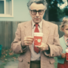 In this image, we see a man and a little girl standing together, both holding cups of coffee. The man is wearing a suit and glasses, while the little girl is wearing a casual outfit. The man''s face shows a mature age, possibly around 55 years old, and he is holding his coffee cup with a heart design on it. The little girl looks to be around 6 years old. The man''s attire suggests a formal setting, possibly a business or professional environment. The background shows a trash bin can and a belt, adding to the scene''s context.