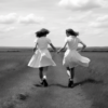 In this black and white photo, we see two women dressed in white dresses walking hand in hand on a dirt road. The women are walking confidently, showcasing a sense of camaraderie and unity. One woman is wearing sneakers, while the other is wearing other shoes. The setting is serene, with a vast field surrounding the dirt road. The contrast between the white dresses and the dark road creates a striking visual impact. The image exudes a sense of friendship, freedom, and togetherness as the women walk together in harmony.