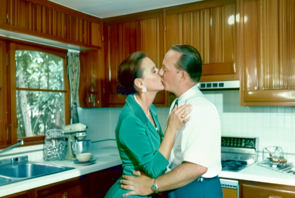 In this image, we see a romantic scene unfolding in a cozy kitchen. A man and a woman are sharing a tender kiss near a kitchen island. The woman is wearing a green shirt, and the man has short hair. The kitchen is well-equipped with wooden cabinets, a sink, a microwave, and a gas stove. There are various kitchen utensils and appliances scattered around, such as plates, bowls, a tea pot, and a bottle. The colors in the kitchen are primarily earthy tones like brown and green, creating a warm and inviting atmosphere. The focus is on the couple, capturing a moment of intimacy amidst the hustle and bustle of a busy kitchen.