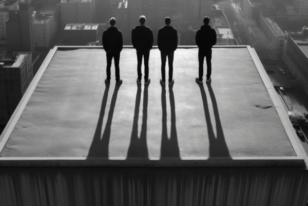 Three men are standing on a roof in the city, with one man wearing sneakers, another man wearing other shoes, and the third man wearing sneakers as well. The men are positioned in a row, with the first man on the left side of the image, the second man in the middle, and the third man on the right side. The men are all facing forward, looking towards the cityscape in the distance. The image is in black and white, adding a dramatic and urban feel to the scene. The men cast long shadows on the rooftop, emphasizing the height of their location.