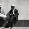 Two men are depicted in a black and white photograph, sitting on a bench in a public area. Both men are elegantly dressed, one wearing a suit and tie with a hat, and the other wearing a suit. They are engrossed in reading newspapers, with one man holding a book as well. The scene is captured in a timeless manner, showcasing a classic and sophisticated style. The setting includes a wooden bench, with the men''s leather shoes visible on the floor. The image exudes a sense of relaxation and intellectual engagement as the men enjoy their reading material.